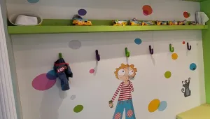 Vestiaire de la crèche : étagère verte, patères colorées et mur décoré d'un personnage