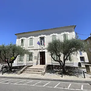 Façade d'une mairie pavoisée, précédée d'oliviers, sous un ciel bleu